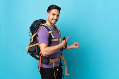 Sırt çantası ve yürüyüş direkleri olan beyaz bir adam mavi arka planda izole edilmiş.