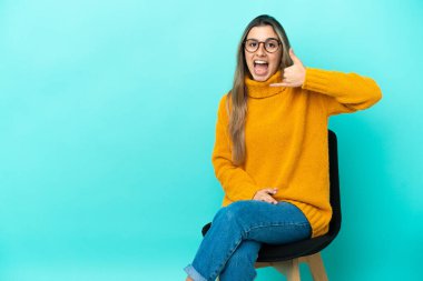 Mavi arka planda izole edilmiş bir sandalyede oturan beyaz bir kadın telefon jesti yapıyor. Bana geri dönüş işareti ver.