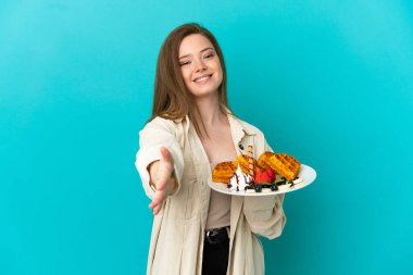 İzole edilmiş mavi arka planda waffle tutan genç kız iyi bir anlaşma için el sıkışıyor.