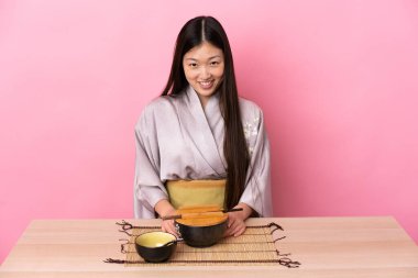 Genç Çinli kız kimono giyiyor ve pembe arka planda bir masada erişte yiyor.
