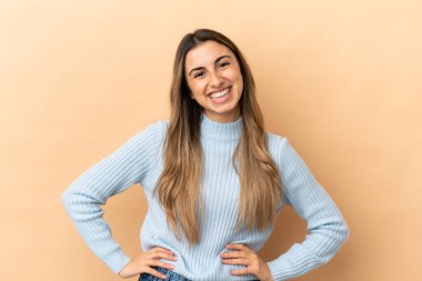 Beyaz tenli genç bir kadın bej arka planda izole edilmiş. Kolları kalçasında ve gülümsüyor.