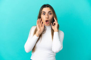 Mavi arka planda izole edilmiş, şaşırmış ve şok olmuş bir yüz ifadesiyle cep telefonu kullanan beyaz bir kadın.
