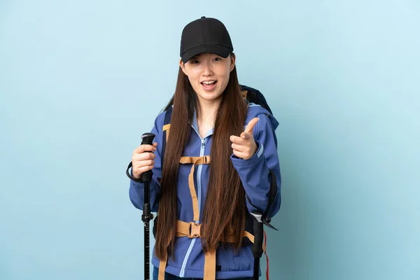 一个年轻的中国姑娘 背着背包 带着远足的杆子 背着蓝色的孤零零的背景 指着前面笑着 — 图库照片