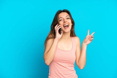 Mavi arka planda izole edilmiş cep telefonu kullanan genç beyaz kadın gülümsüyor ve zafer işareti gösteriyor.