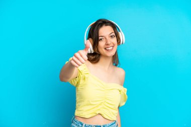 Genç beyaz kadın mavi arka planda izole edilmiş müzik dinliyor ve başparmağını kaldırıyor.