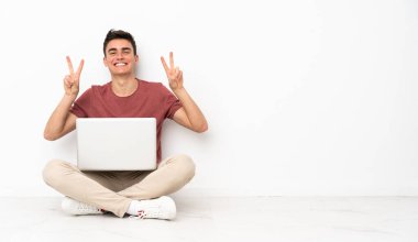 Flor 'da dizüstü bilgisayarıyla oturan genç adam iki eliyle zafer işareti gösteriyor.