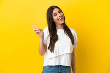 Sarı arka planda izole edilmiş genç beyaz kadın gülümsüyor ve zafer işareti gösteriyor.