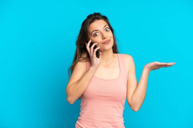 Mavi arka planda izole edilmiş cep telefonu kullanan genç beyaz kadın ellerini kaldırırken şüpheye düşüyor