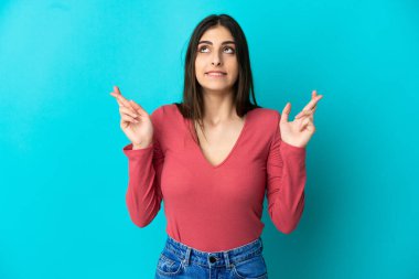 Mavi arka planda izole edilmiş genç beyaz kadın parmaklarını çapraz yapıp en iyisini diliyor.