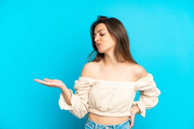 Mavi arka planda izole edilmiş genç beyaz bir kadın. Kopyalama alanını şüphe ile tutuyor.