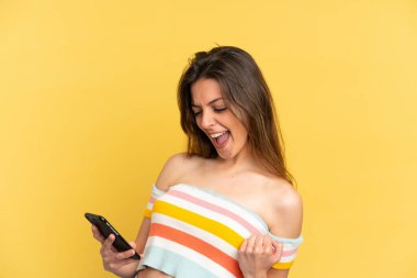 Sarı arka planda izole edilmiş genç beyaz kadın cep telefonu kullanıyor ve zafer jesti yapıyor.