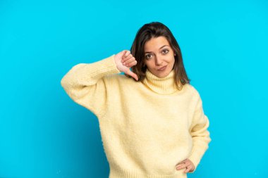 Mavi arka planda izole edilmiş genç beyaz kadın olumsuz ifadeyle baş parmağını aşağı gösteriyor.
