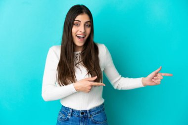 Mavi arka planda izole edilmiş genç beyaz kadın şaşırmış ve işaret ediyor.