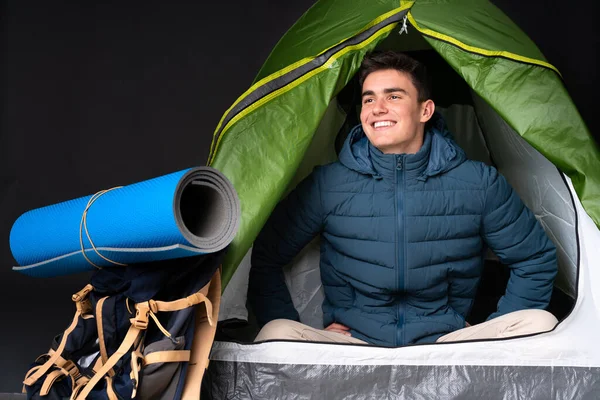 Teenager caucasian man inside a camping green tent isolated on black background posing with arms at hip and smiling
