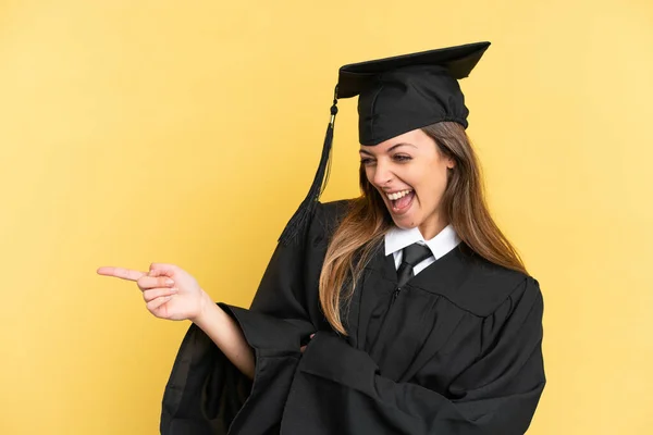 Young university graduate isolated on yellow background pointing finger to the side and presenting a product