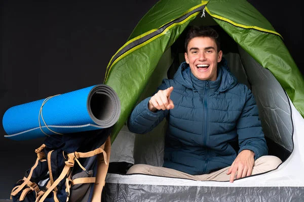 Teenager caucasian man inside a camping green tent isolated on black background surprised and pointing front