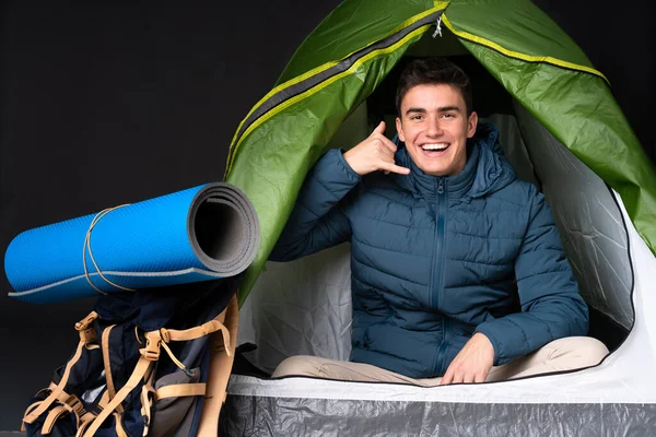 Teenager caucasian man inside a camping green tent isolated on black background making phone gesture. Call me back sign