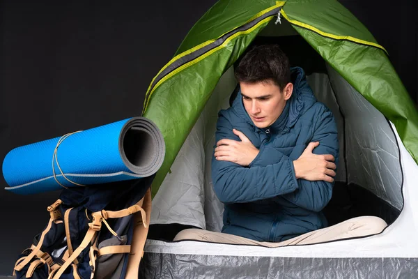 Teenager caucasian man inside a camping green tent isolated on black background freezing