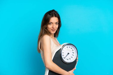 Genç beyaz kadın, mavi arka planda, tartı makinesiyle izole edilmiş.
