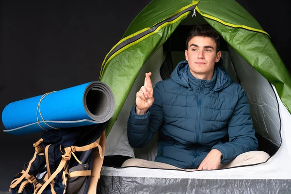 Teenager caucasian man inside a camping green tent isolated on black background with fingers crossing and wishing the best