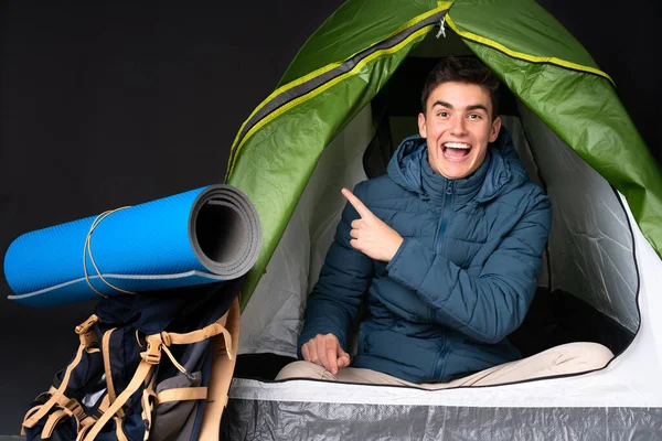 Teenager caucasian man inside a camping green tent isolated on black background surprised and pointing side