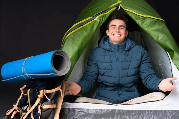 Teenager caucasian man inside a camping green tent isolated on black background smiling