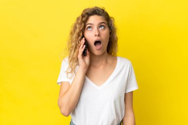 Sarı arka planda izole edilmiş cep telefonu kullanan genç sarışın kadın yukarı bakıyor ve şaşırmış bir ifadeyle