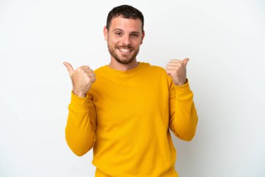 Beyaz arka planda izole edilmiş genç Brezilyalı bir adam. Baş parmakları havada ve gülümsüyor.