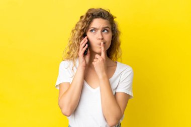 Sarı arka planda izole edilmiş cep telefonu kullanan genç sarışın kadın yukarı bakarken şüphe duyuyor.