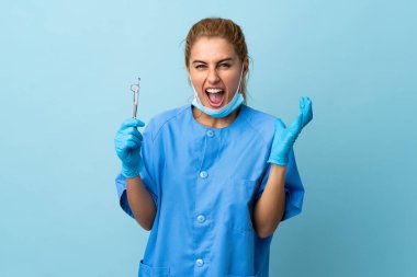 İzole edilmiş mavi arka planda aletleri tutan genç dişçi kadın mutsuz ve hayal kırıklığına uğramış.