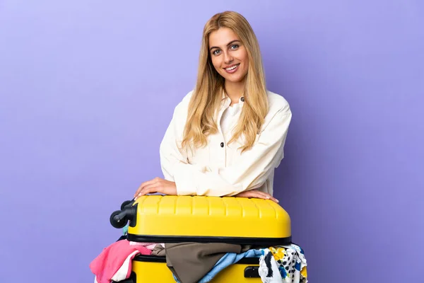 Young Uruguayan blonde woman with a suitcase full of clothes over isolated purple wall laughing