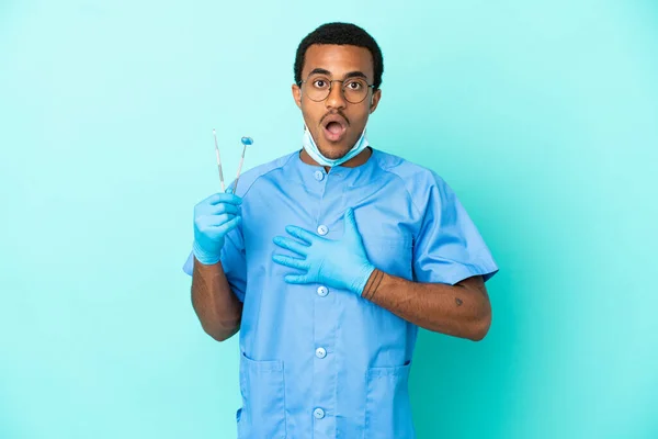 African American dentist holding tools over isolated blue background surprised and shocked while looking right