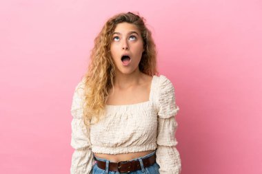 Pembe arka planda izole edilmiş genç sarışın kadın yukarı bakıyor ve şaşırmış bir ifadeyle