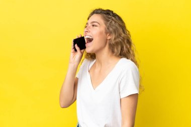 Sarı arka planda izole edilmiş cep telefonu kullanan genç sarışın kadın yan pozisyonda gülüyor.