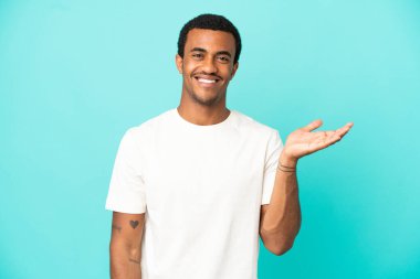 İzole edilmiş mavi arka planda oturan Afro-Amerikalı yakışıklı bir adam bir ilan vermek için avucunda hayali bir şey tutuyor.