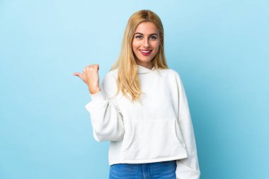 Genç Uruguaylı sarışın kadın izole edilmiş mavi arka planda bir ürün sunmak için kenarı işaret ediyor.