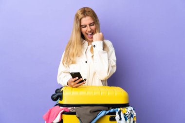 Genç Uruguaylı sarışın kadın bir bavul dolusu elbisesi ile izole edilmiş mor duvarın üzerinde telefon zafer pozisyonunda.