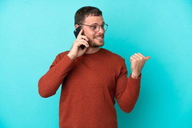 Mavi arka planda izole edilmiş cep telefonu kullanan genç Brezilyalı bir adam bir ürün sunmak için kenarı işaret ediyor