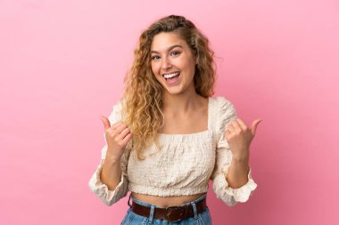 Pembe arka planda izole edilmiş genç sarışın kadın. Baş parmakları havada ve gülümsüyor.