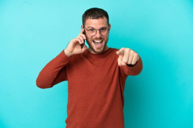 Mavi arka planda izole edilmiş cep telefonu kullanan genç Brezilyalı adam şaşırmış ve ön tarafı işaret ediyor.