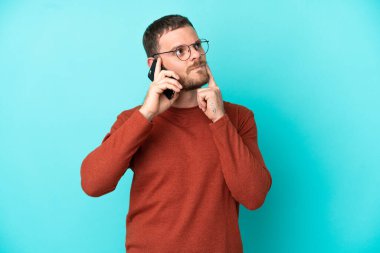 Mavi arka planda izole edilmiş cep telefonu kullanan genç Brezilyalı adam yukarı bakarken şüphe duyuyor