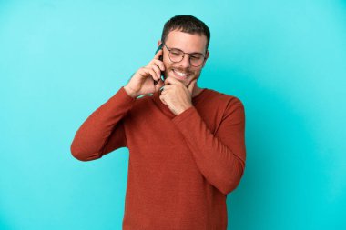 Mavi arka planda izole edilmiş cep telefonu kullanan Brezilyalı genç adam yana bakıyor ve gülümsüyor.