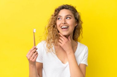 Sarı arka planda dişlerini fırçalayan beyaz bir kadın yan tarafa bakıyor ve gülümsüyor.