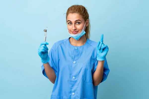 Młoda Kobieta Dentysta Trzyma Narzędzia Odizolowanym Niebieskim Tle Wskazując Palcem — Zdjęcie stockowe