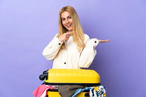 若いですウルグアイブロンド女性とともにスーツケースフルの服上の隔離された紫色の壁保持コピースペース想像上のザ手のひらに挿入する広告 — ストック写真
