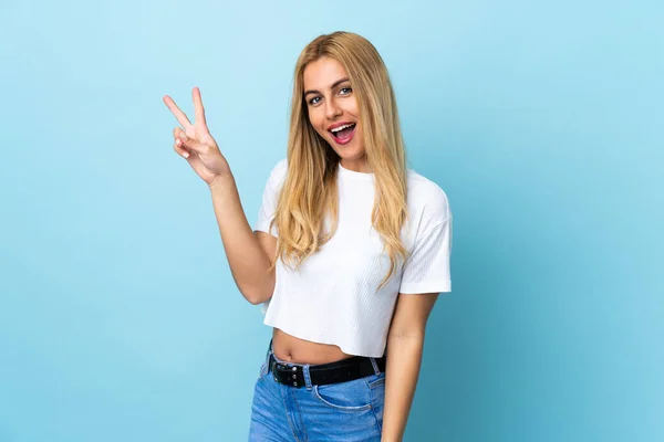Молода Уругвайська Блондинка Над Синім Фоном Посміхається Показує Знак Перемоги — стокове фото