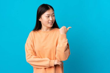 İzole edilmiş mavi arka planda genç bir Çinli kız bir ürünü sunmak için kenarı işaret ediyor.