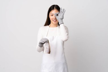 Çinli balıkçı önlük takıyor ve beyaz arka planda çiğ balık tutuyor. Parmaklarıyla işaret veriyor.