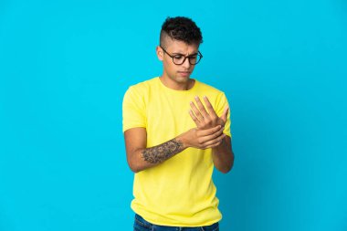 Genç Brezilyalı adam mavi arka planda izole edilmiş, ellerinde acıdan muzdarip.