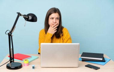 Öğrenci kız mavi arka planda izole bir şekilde evinde çalışıyor. Şüpheleri var ve kafası karışmış bir yüz ifadesi var.